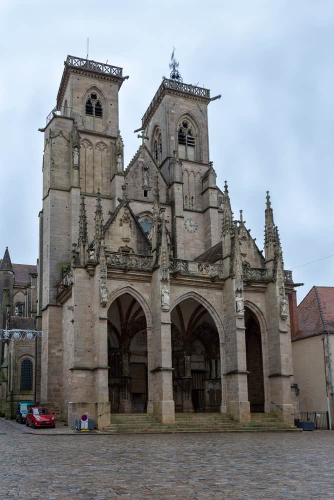 Small square with a huge church in the back and cars parked nearby.