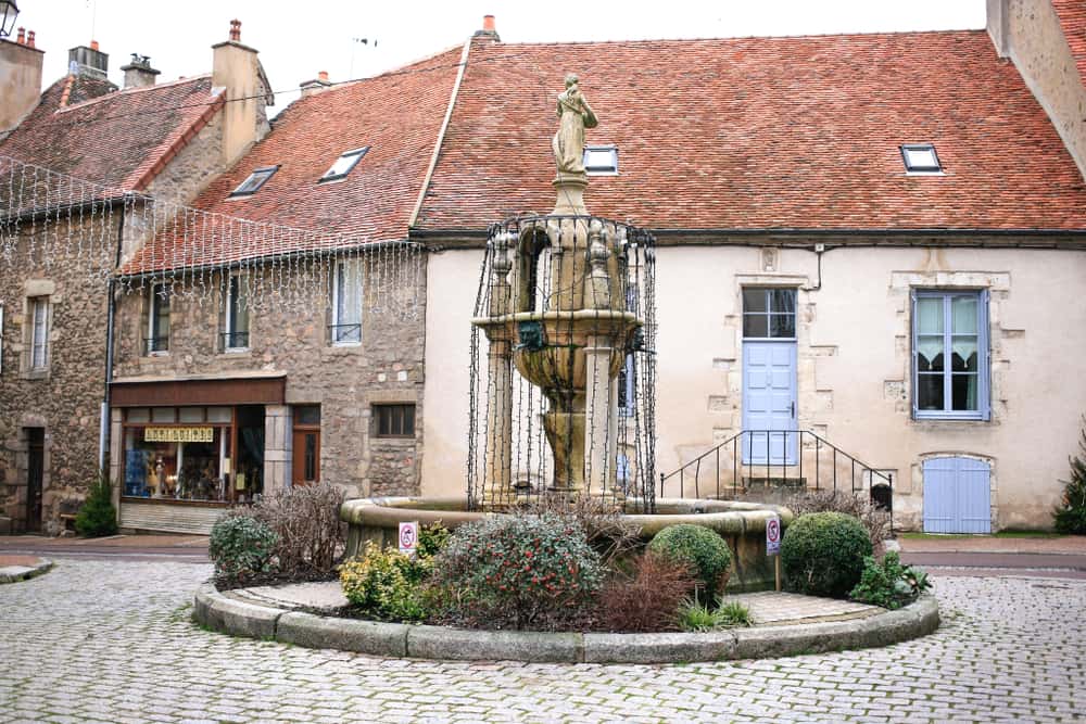 A cozy square in the center of the city of Saulieu in Burgundy, France. There is a big fountain and old houses in the background.