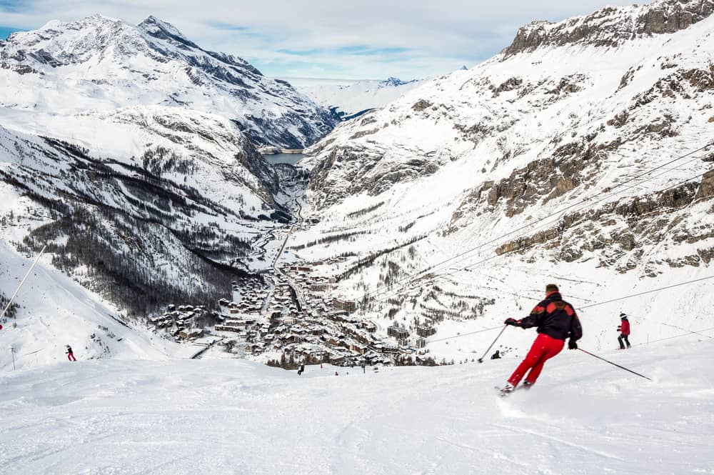 A few skiers going down piste in Val d'Isere, France. Lots of snow and mountains - and a town visible in a valley.