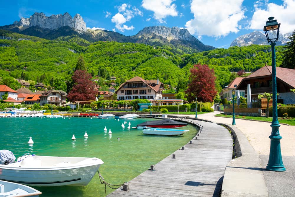 The beautiful town of Talloires in France on a bright and sunny day. A lake and boats with houses and mountains in the background.