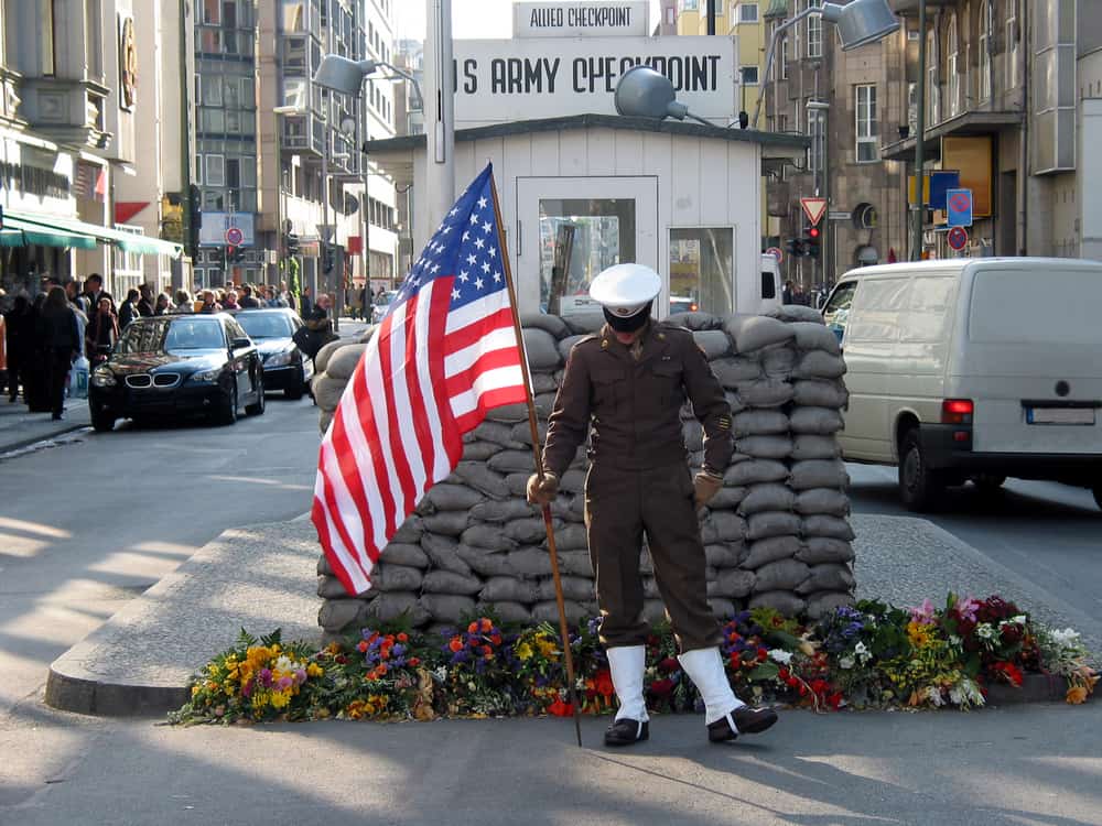 Replica of Checkpoint Charlie in the middle of a busy street in Berlin. In front a US soldier holding the American flag.